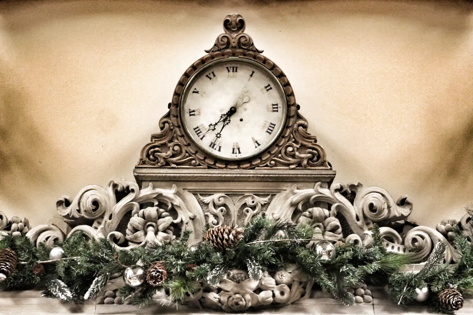 An ornate carved clock on a marble mantel above an evergreen garland with pinecones and silver ornaments