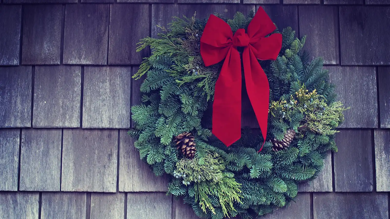 A Christmas wreath of evergreen branches and pinecones with a red bow, hung on grey wooden shingles