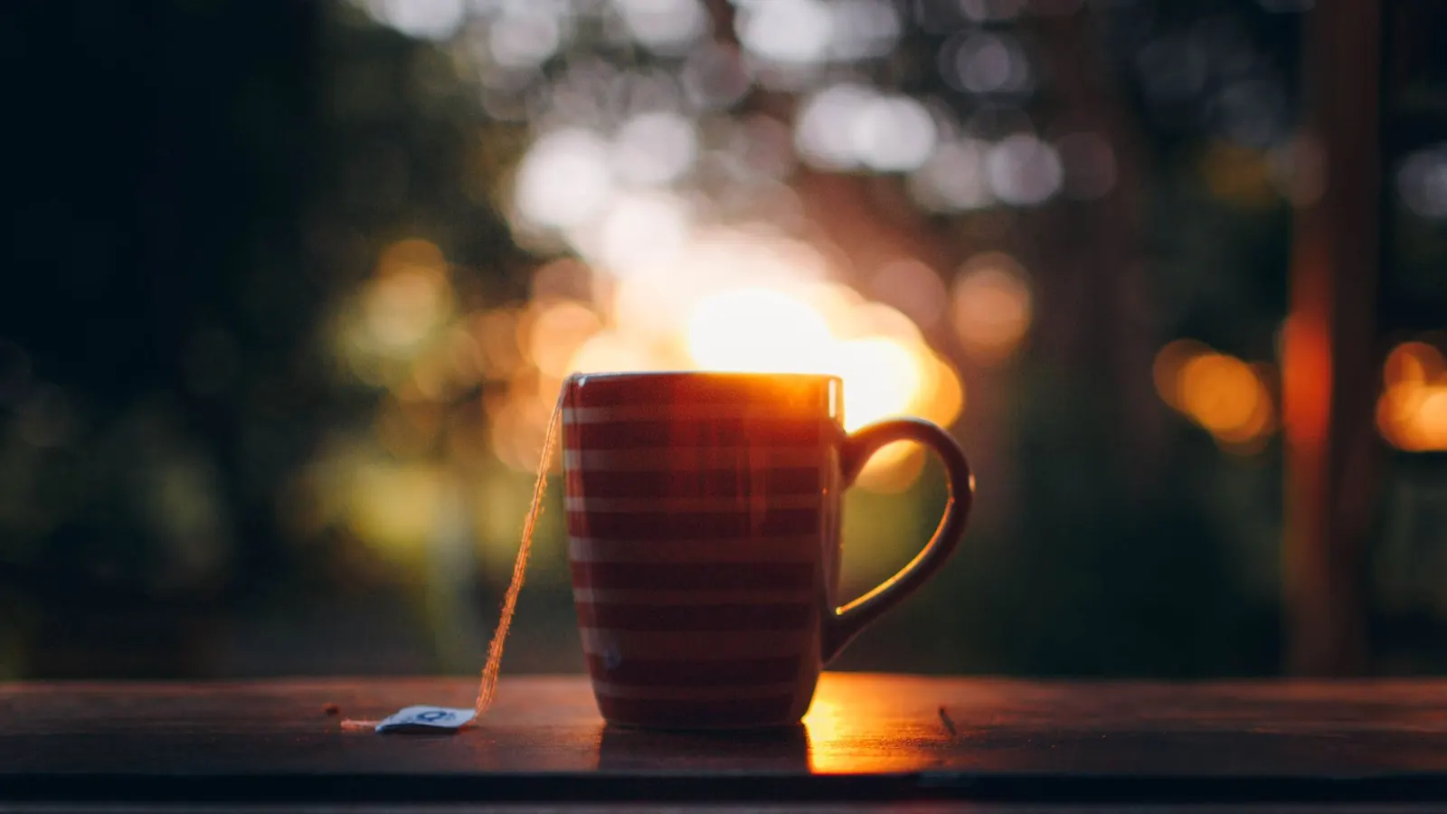 A striped mug of hot tea with a tea bag string hanging over the side, on a wooden windowsill at sunset