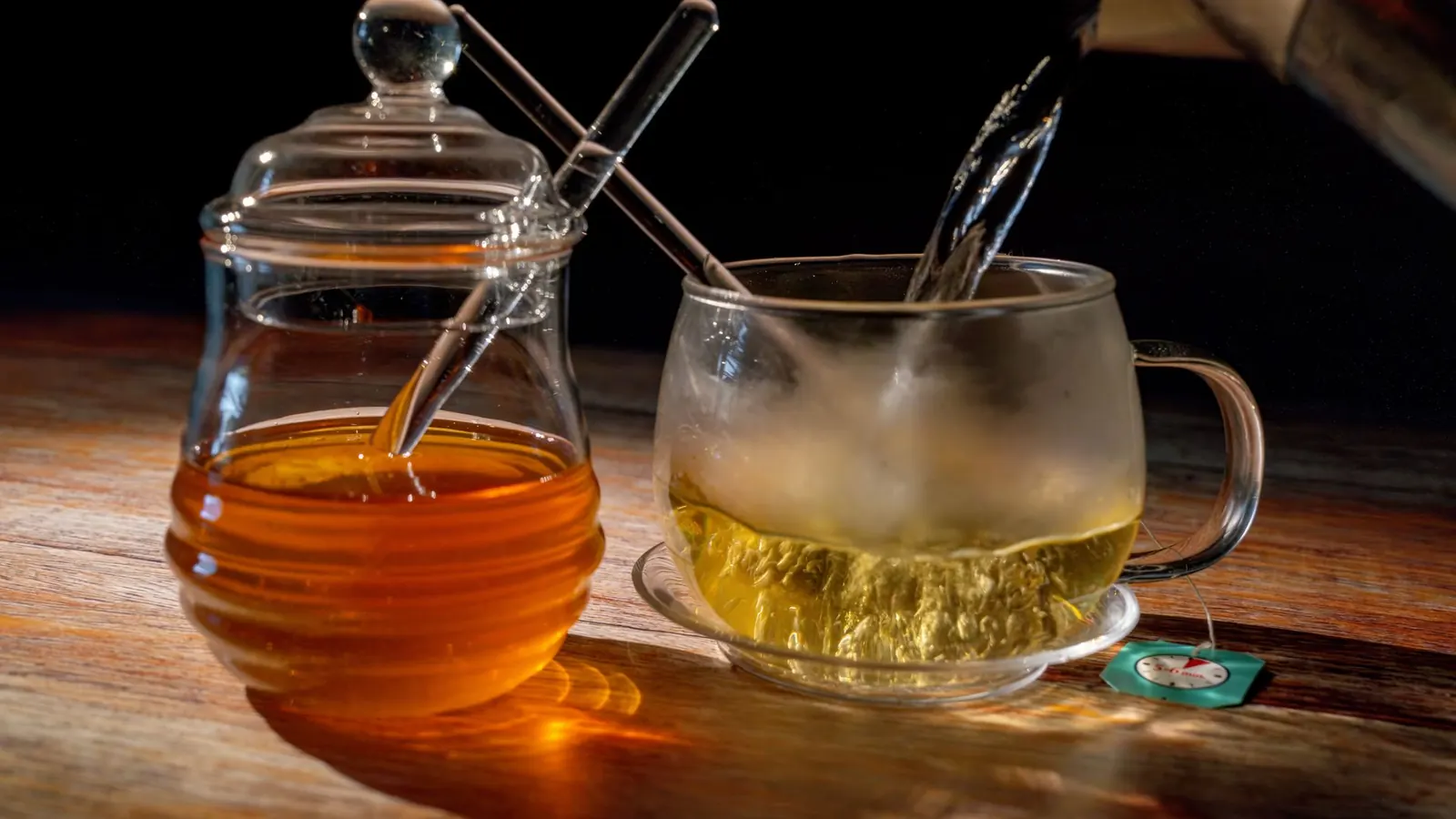 Hot tea being poured into a glass cup on a saucer beside a glass jar of honey on a wooden table