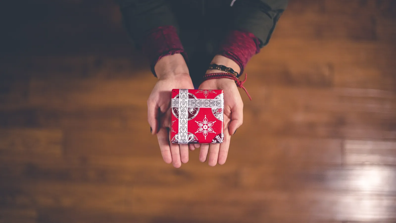 Two open hands holding a small gift wrapped in red paper with a white snowflake pattern