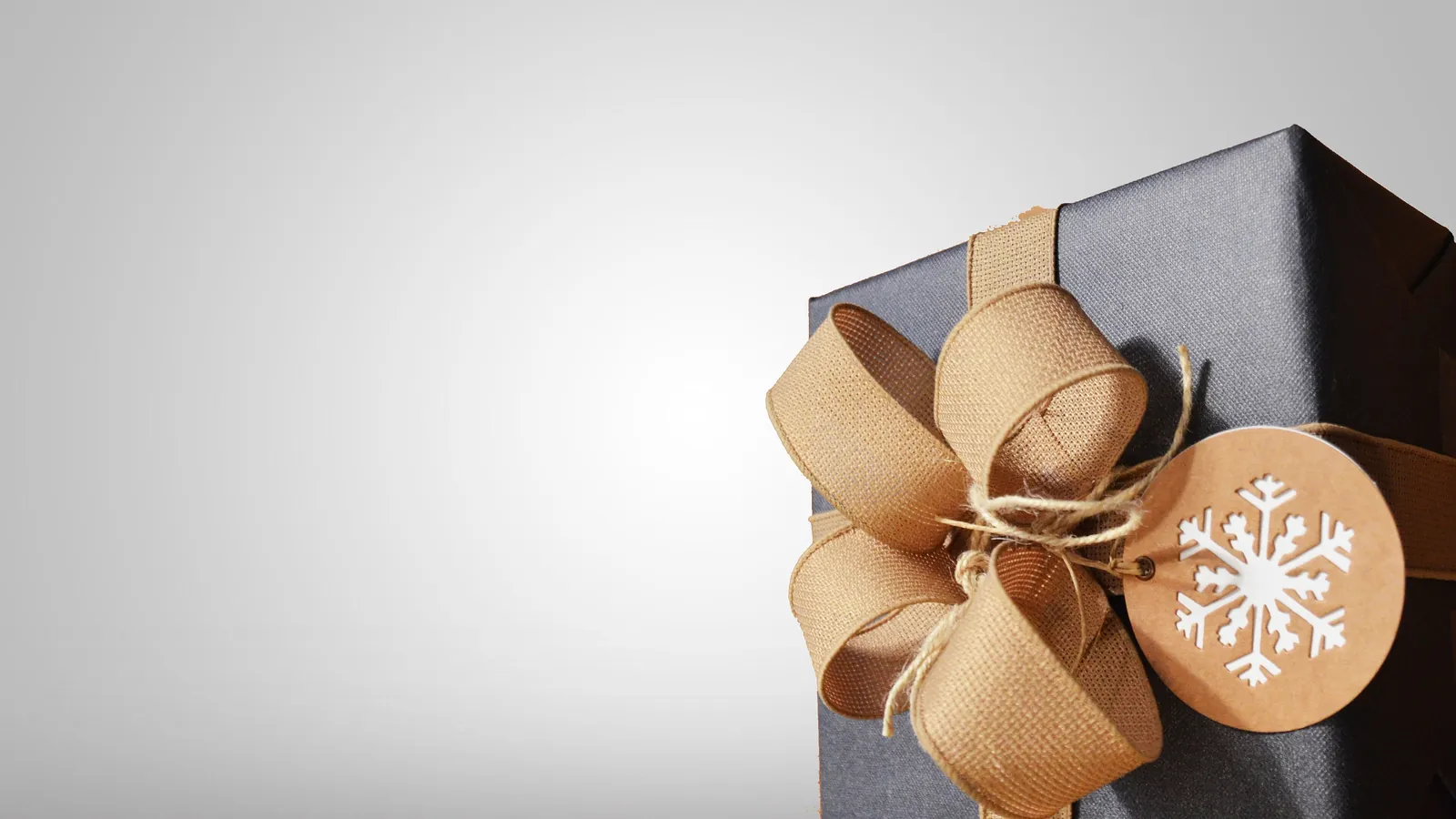 A dark gift box tied with a wide burlap ribbon bow and a round kraft tag printed with a white snowflake