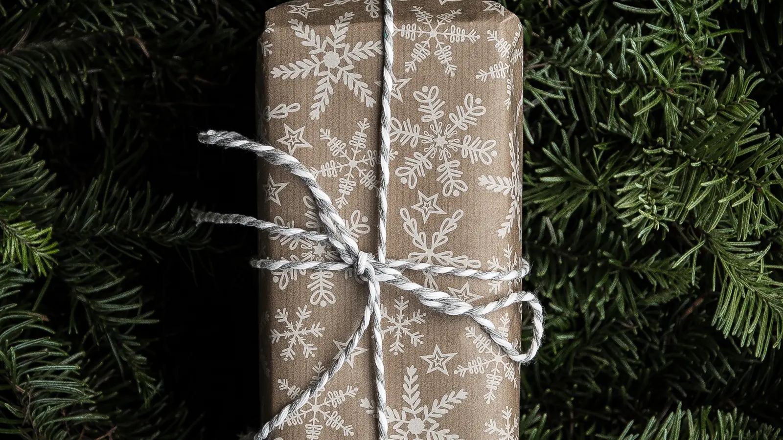 A gift wrapped in brown paper with a white snowflake print and white twine, lying on fir branches