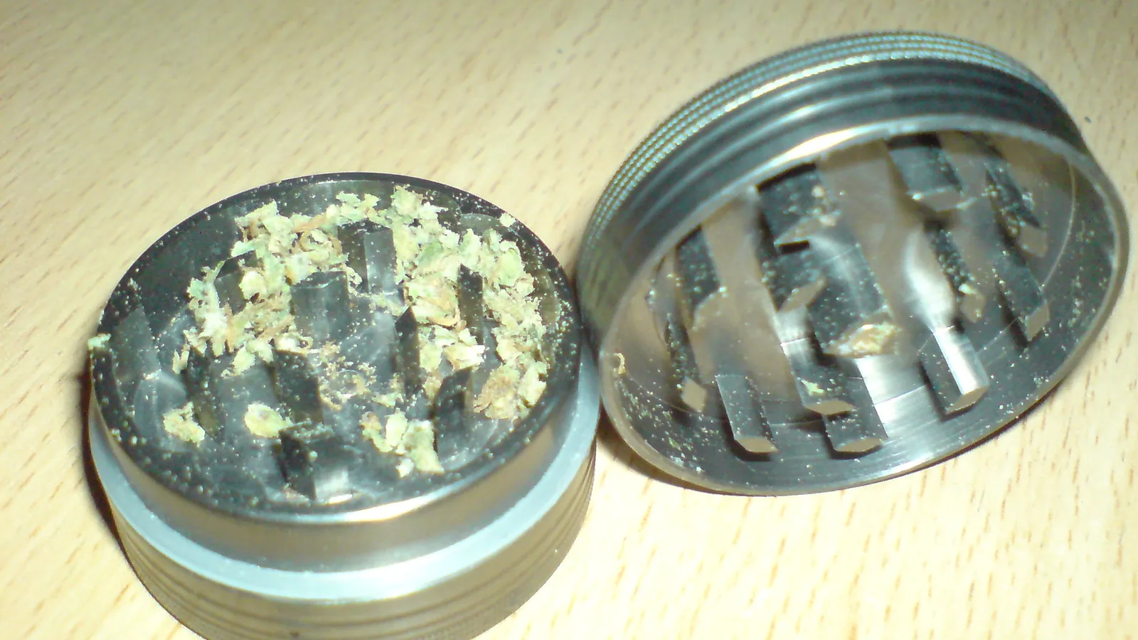 An open metal herb grinder on a wooden table with a little ground cannabis between its teeth